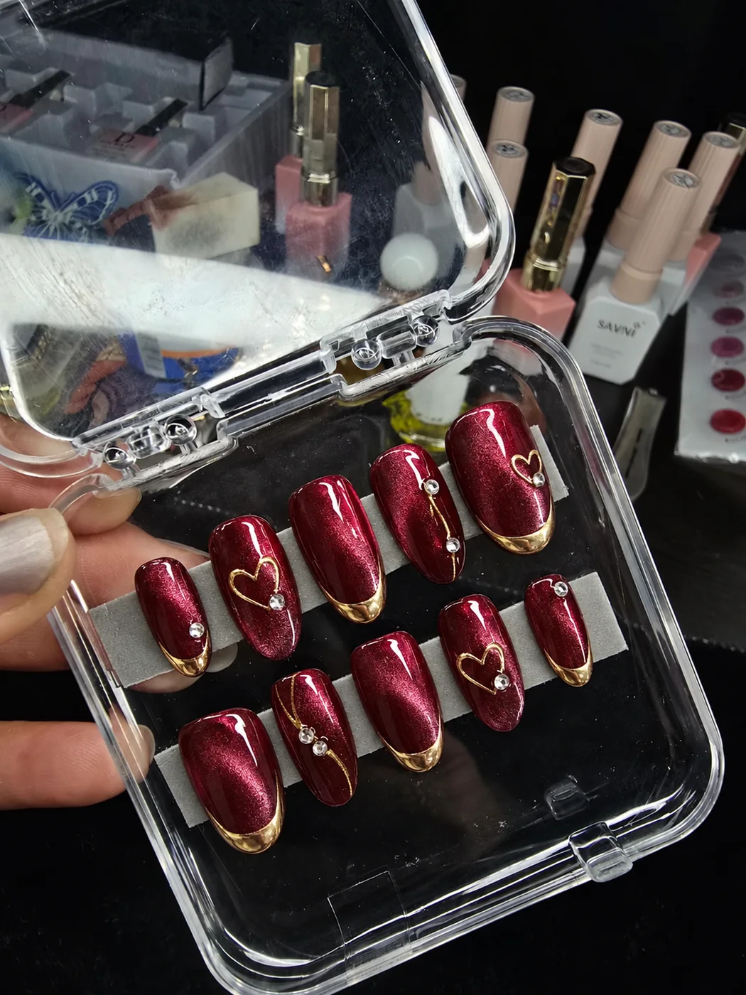 Burgundy press-on nails with gold details arranged in a clear tray